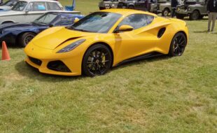 Yellow Lotus Emira in front of other Lotus cars and Land Rovers on a grassy field